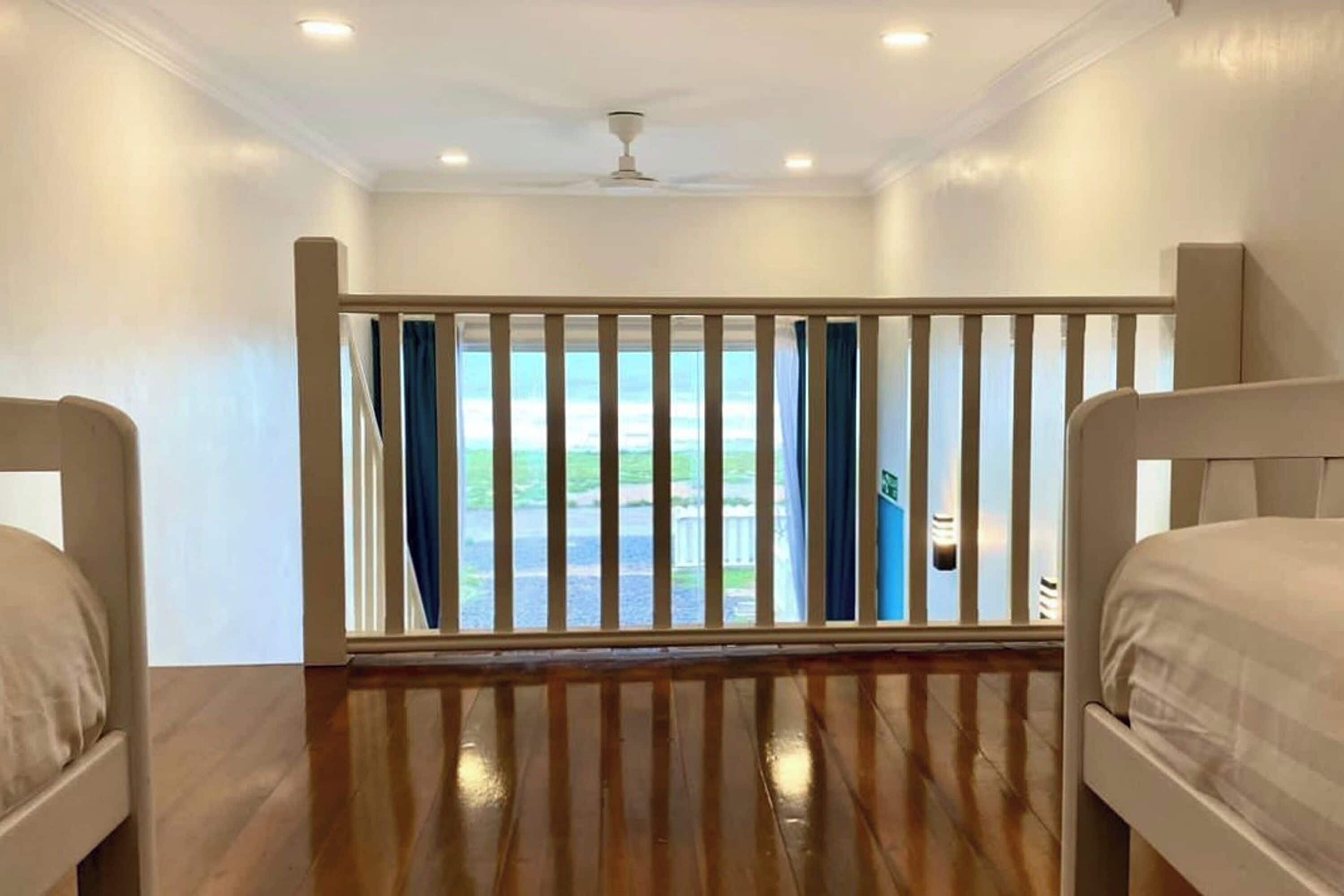 A room with two beds, a wooden floor, a balcony with a railing, and a view of the outdoors.