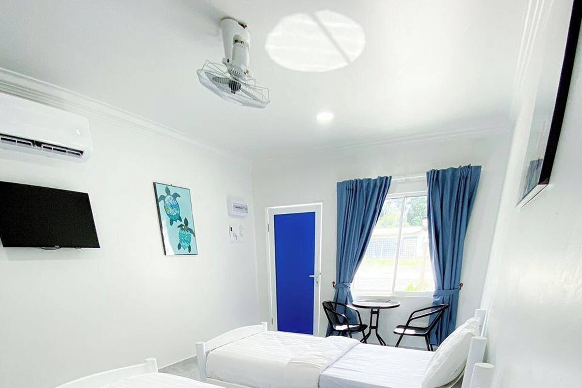 Habitación de hotel con dos camas individuales, una mesita pequeña, sillas, televisor y ventilador de techo.