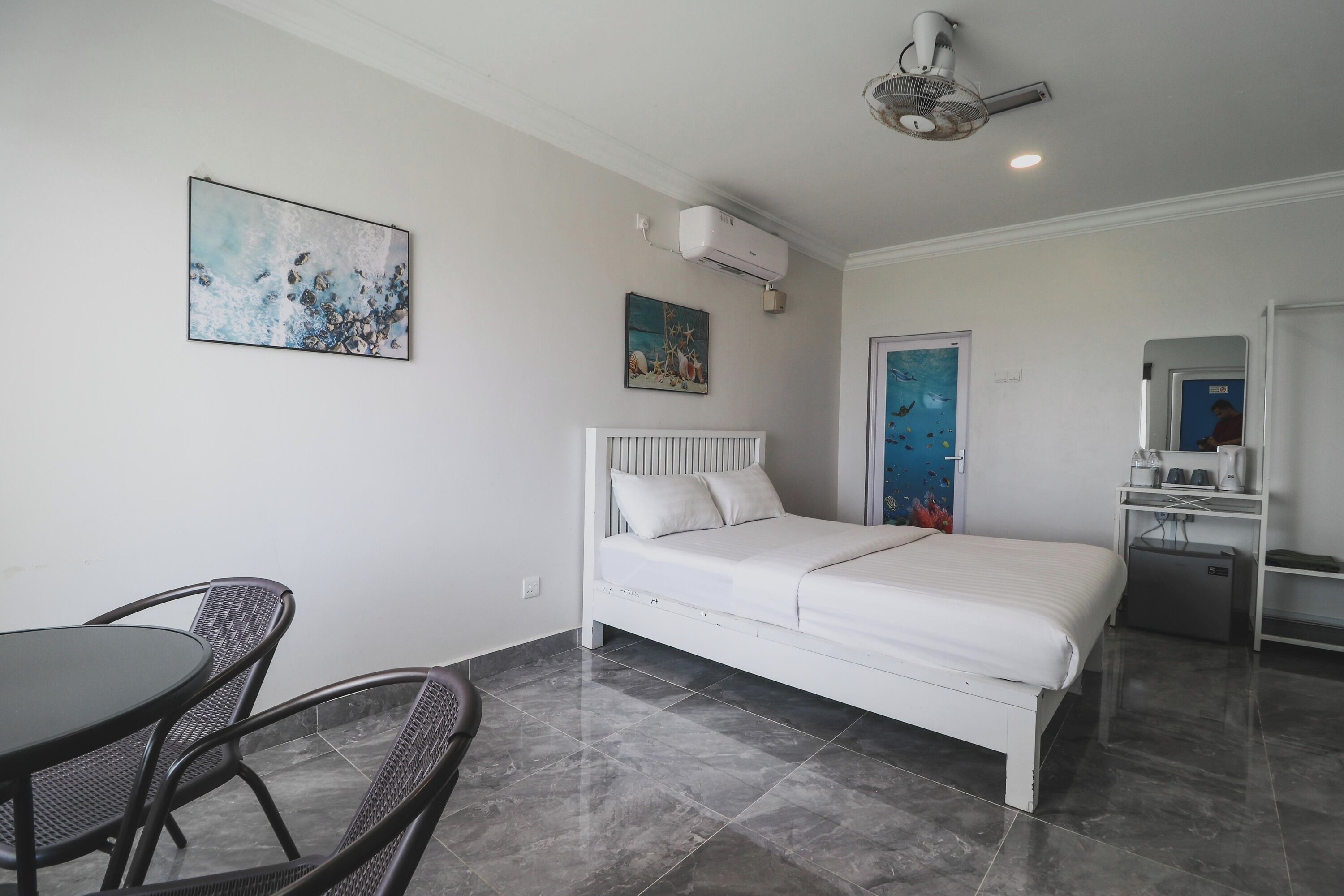 Un dormitorio con cama, una mesita, dos sillas, un ventilador de techo, aire acondicionado y una puerta con un cartel.