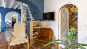 Una habitación con pared de ladrillo blanco, un televisor montado en la pared, una silla de madera y un sillón con cojines, y una escalera que lleva a otra zona.
