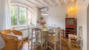 Un comedor con una mesa redonda, sillas, un sillón de mimbre, un armario y una ventana con cortinas.
