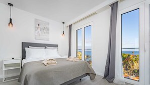 A modern bedroom with a large bed, a nightstand, a wall-mounted light, and a view of the sea. - Where Serenity Meets Blue Horizons.  (Câmara de Lobos)