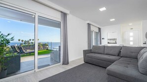 A modern living room with a grey sofa, a large sliding glass door leading to a balcony with a view of the sea, and a kitchen area visible in the background. - Where Serenity Meets Blue Horizons.  (Câmara de Lobos)