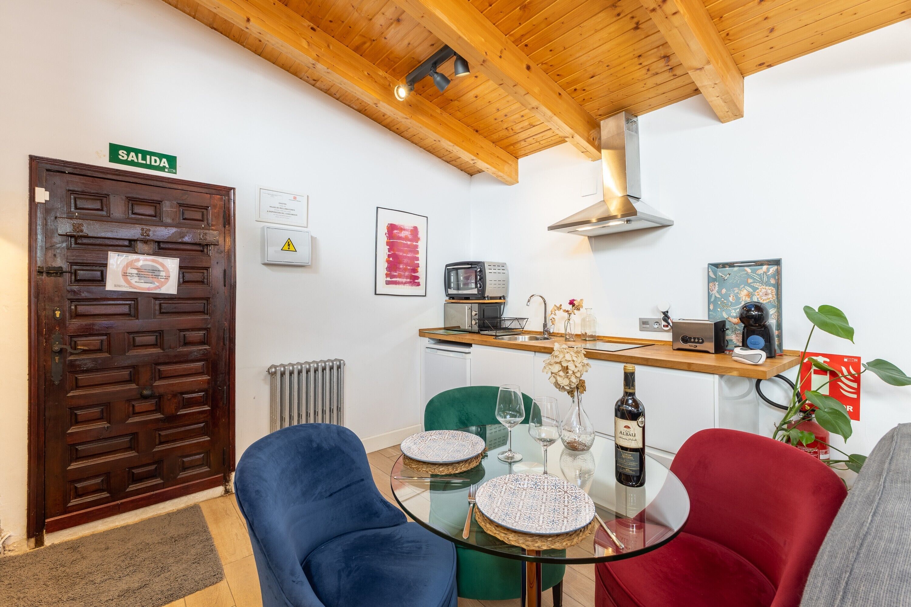 A room with a wooden ceiling, a door marked 'SALIDA,' a small kitchenette, and a glass table with chairs.