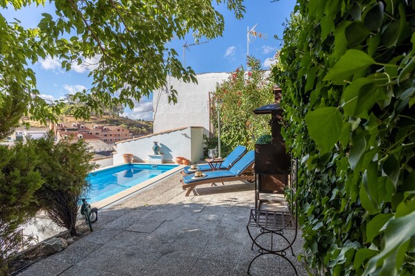 Zona de piscina con sillones reclinables, una pérgola y vista a edificios al fondo.