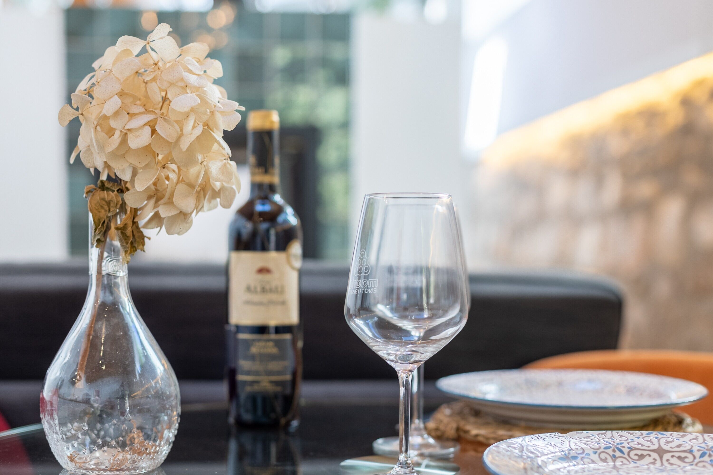 Una mesa con un jarrón de vidrio transparente con flores blancas, una botella de vino y una copa de vino vacía.