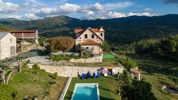 Un edificio de piedra con techo de tejas rojas, una piscina y montañas al fondo.