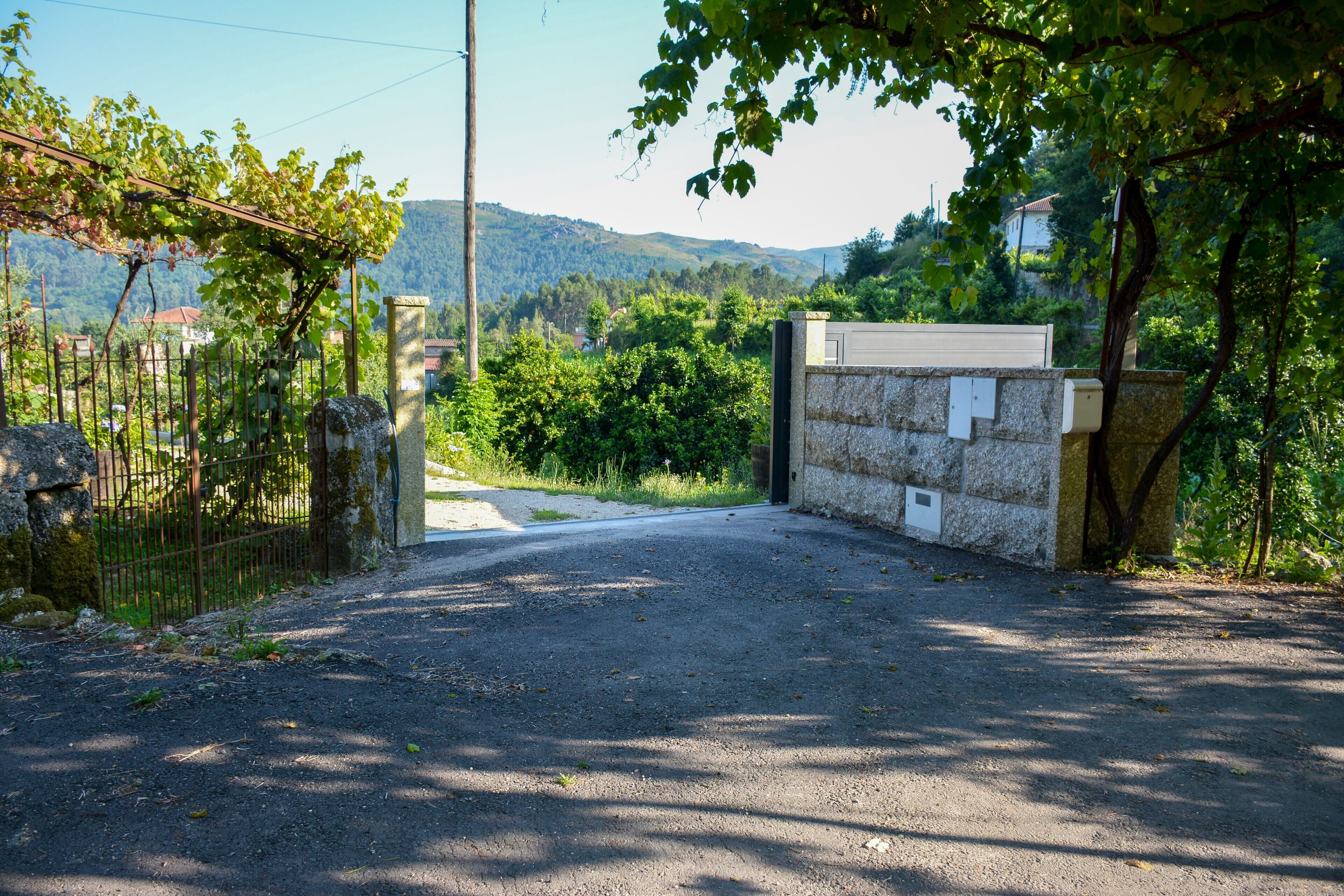 Une allée pavée mène à une entrée fermée par un portail, elle-même bordée d’un mur de pierre et d’un portail en métal, le tout étant entouré de verdure et avec des collines en arrière-plan.