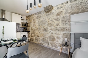 A modern kitchen with a stone wall, a dining area with a table and chairs, and a bedroom with a bed and bedside table.