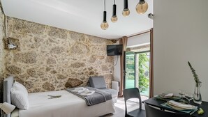 A bedroom with a stone wall, a bed, a TV, a dining table with chairs, and a view of greenery outside. - Studio Apartment 'Quinta Do Bairro -' with Mountain View, Shared Pool and Wi-Fi (Rossas)