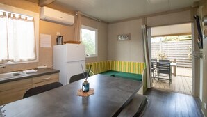 A compact kitchen with a dining area, a window with curtains, a refrigerator, and a view of the outdoor patio. - Fully Equipped Chalet V Near the Sea with Shared Pool, Private Terrace, and Air Conditioning (Pianottoli-Caldarello)