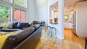 Amplia sala de estar con sofá de cuero, comedor y cocina con alacenas blancas y piso de madera.