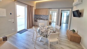 Una sala moderna con una mesa de comedor, sillas blancas, una zona de cocina y un televisor montado en la pared.