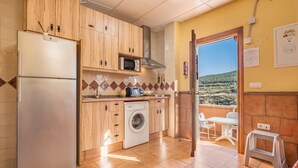 A kitchen with a refrigerator, microwave, washing machine, and a view of a mountainous landscape through an open door. - Apartment 'Tizná' with Mountain View, Shared Terrace and Wi-Fi (Jérez del Maquesado)