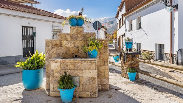 A stone fountain with potted plants, a street with buildings, and a clear sky. - Apartment 'Tizná' with Mountain View, Shared Terrace and Wi-Fi (Jérez del Maquesado)