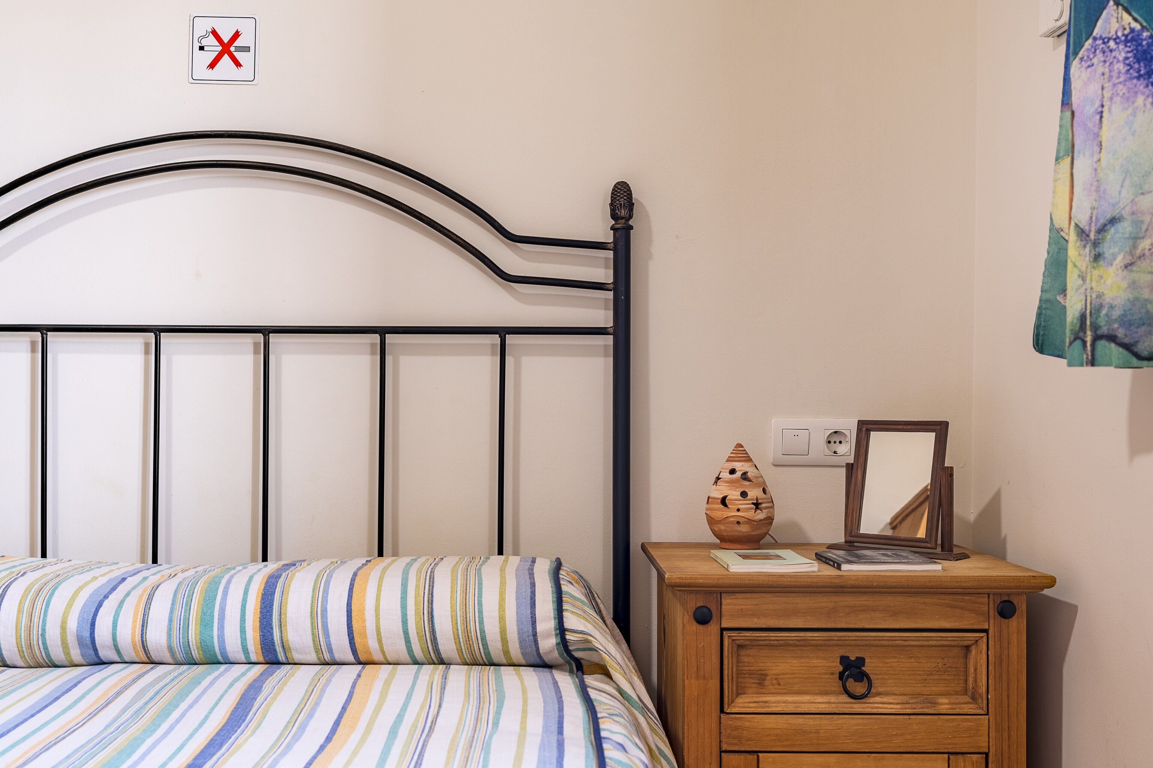 Una cama con colcha a rayas, una mesita de noche de madera con un espejo y un jarrón decorativo, y un interruptor de luz en la pared.