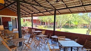 A covered outdoor seating area with wooden tables and chairs, a washroom sign, and a view of a garden. - Rockhill Adventures And Resorts (Roha)