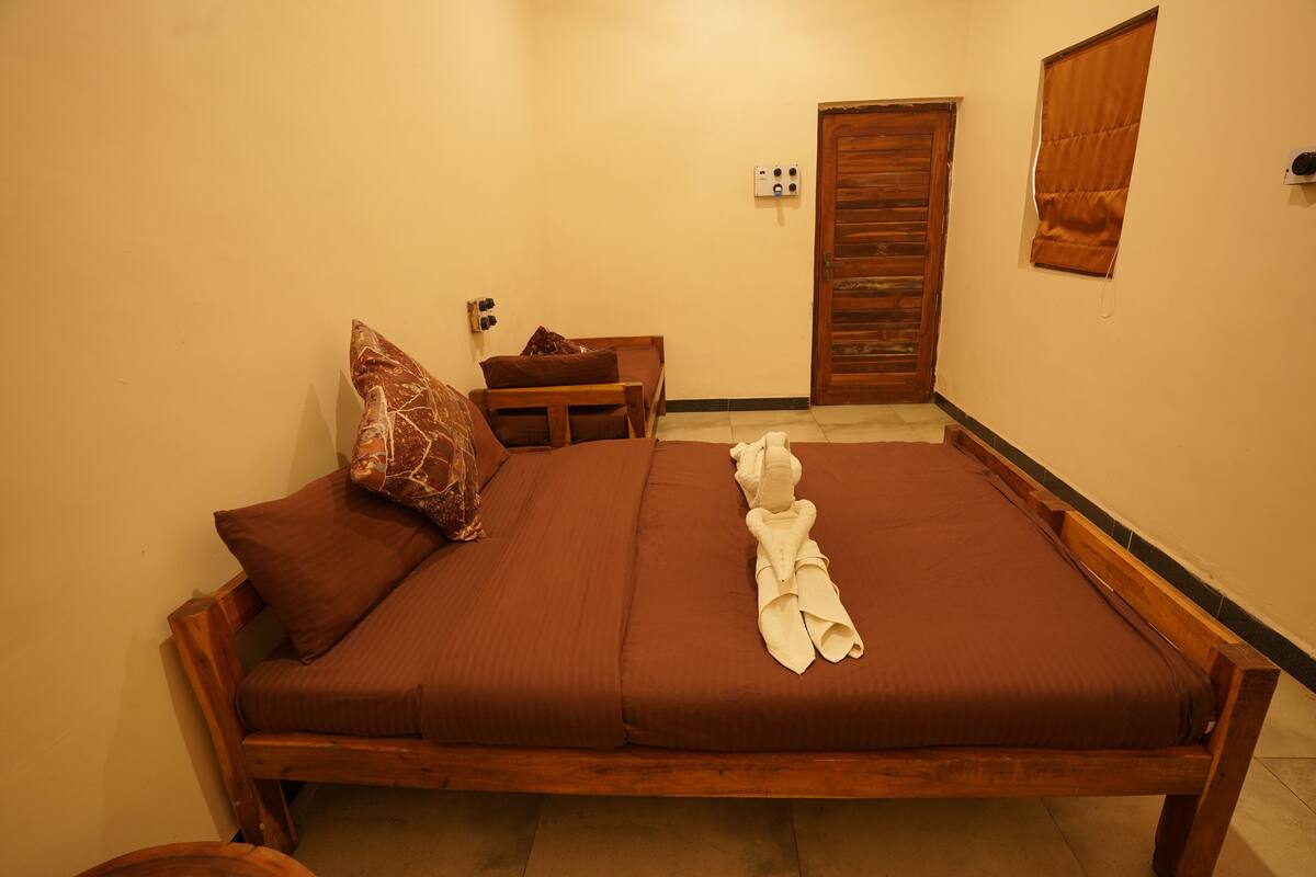 Quarto individual com cama de madeira, roupa de cama marrom, um banco de madeira, uma porta e uma janela com persiana.