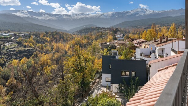 Un paisaje montañoso con picos nevados, árboles de colores otoñales y una mezcla de edificios modernos y tradicionales.
