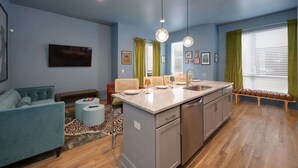 Una sala de estar moderna con un sofá, una mesa de centro y un televisor. Hay una zona de cocina con un fregadero, una encimera y un banco con taburetes.