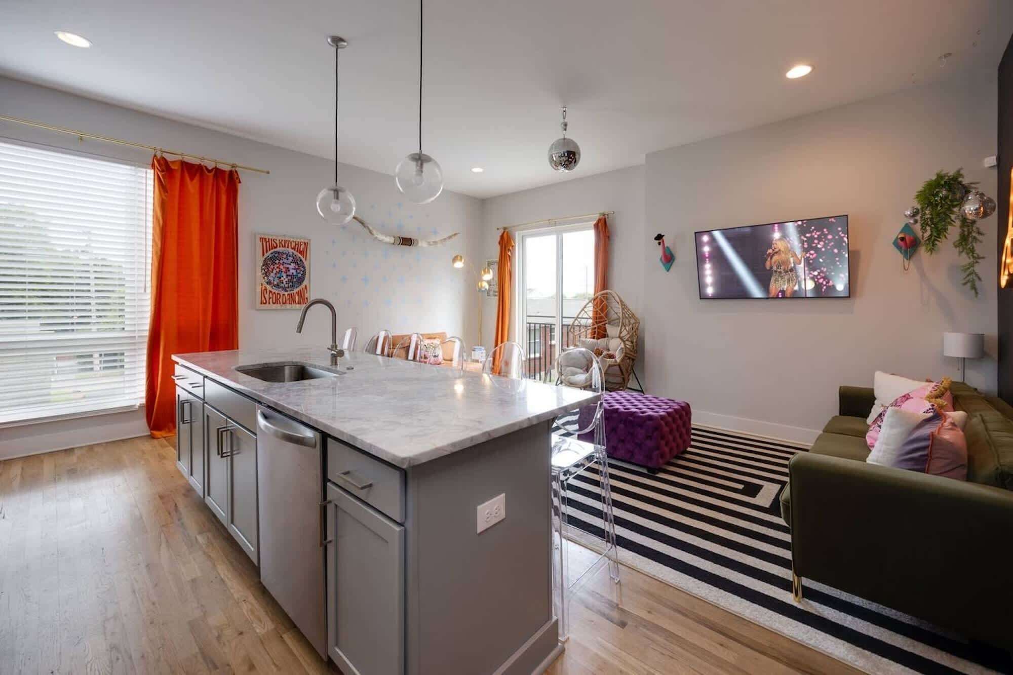 A modern kitchen with a central island, a dining area with chairs, a sofa, and a TV mounted on the wall.