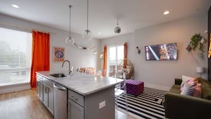 A modern kitchen with a central island, a dining area with chairs, a sofa, and a TV mounted on the wall. - 2BR Near DT — Rooftop Deck + Fire Pit + Sleeps 7 (Nashville)