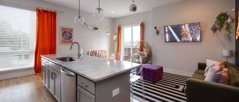 A modern kitchen with a central island, a dining area with chairs, a sofa, and a TV mounted on the wall.