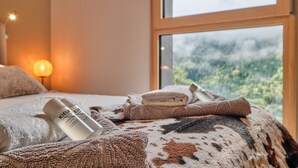 A bed with towels, a bottle of shampoo, and a view of a forest through a window.