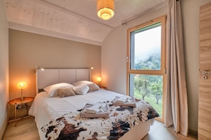 A bedroom with a large bed, bedside lamps, a view of greenery, and a wooden door. - Chalet 'L'Ecrin De Venosc' with Mountain View, Private Terrace and Wi-Fi (Les Deux Alpes)