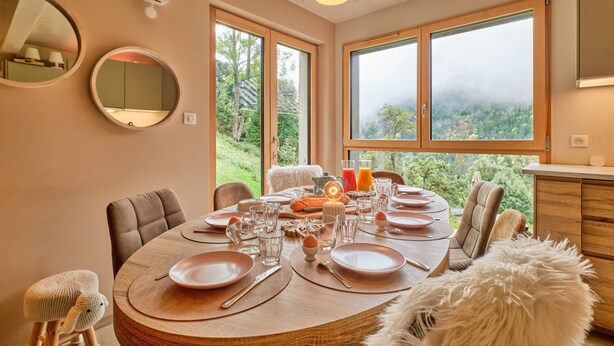 A dining area with a round table set for four, large windows with a view of greenery, and a ceiling light fixture.