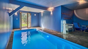 An indoor swimming pool with a view of greenery outside, a bench, and a chair.