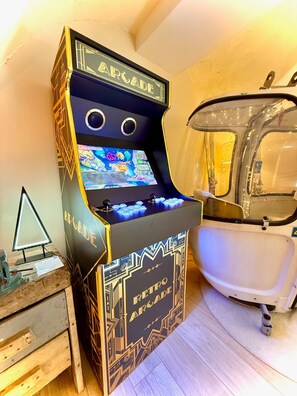 A retro arcade machine with a neon sign, a small table with a lamp, and a bathtub in the background.