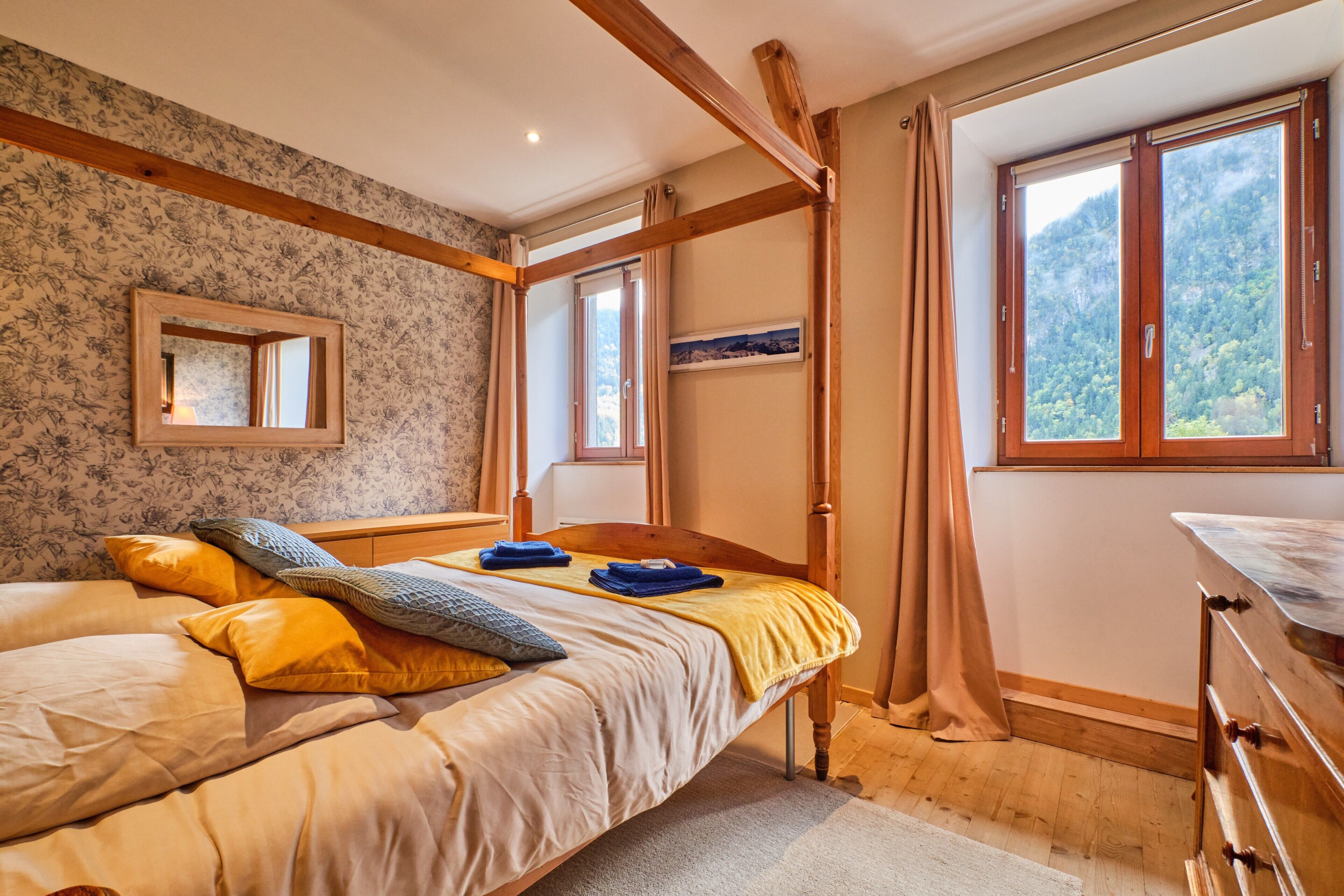 A bedroom with a wooden bed, a mirror, a window with a mountain view, and a dresser.