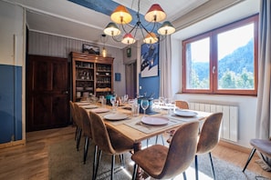 A dining area with a wooden table set for four, surrounded by upholstered chairs, a chandelier, and a view of a mountain through a window.