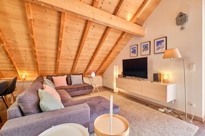 Ein gemütliches Schlafzimmer im Loft-Stil mit schrägem Dach, großem Fenster, Flachbildfernseher, Nachttisch und Lampe.