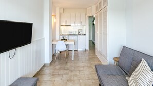 Un espace de vie compact comprenant un coin cuisine, un téléviseur à écran plat fixé au mur, un canapé gris agrémenté d’un coussin à rayures et un petit tabouret en bois.