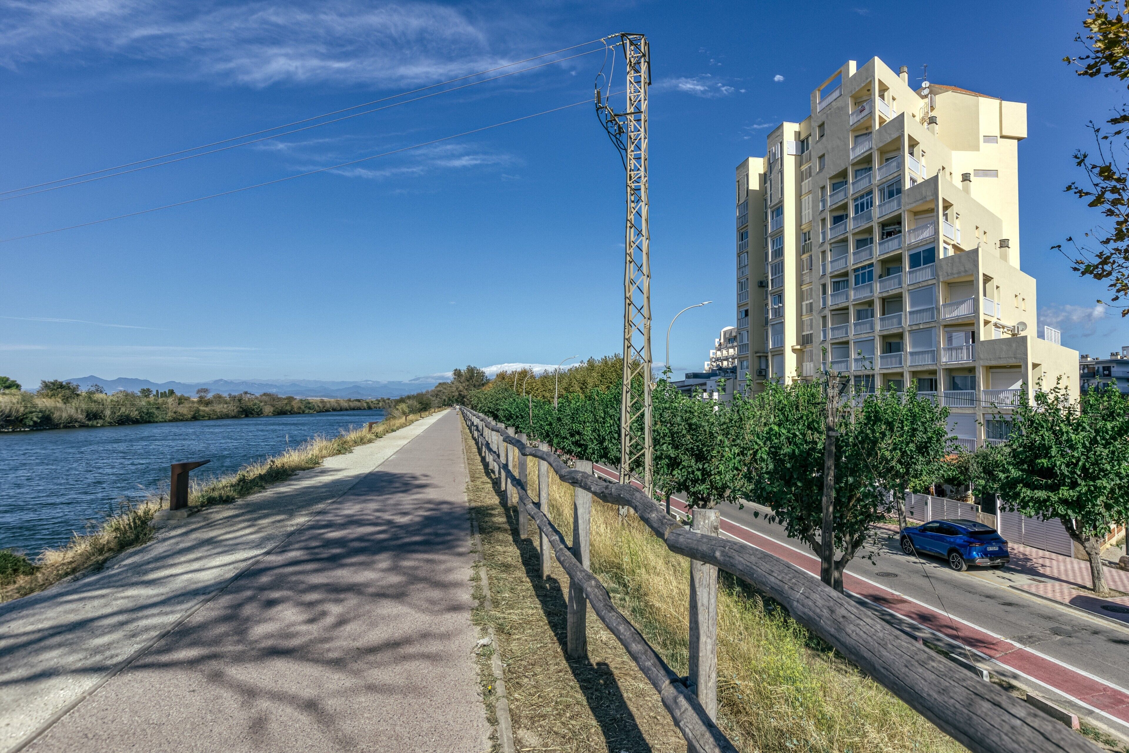 A riverside walkway with a wooden fence, a blue car, and a multi-story building with balconies.
