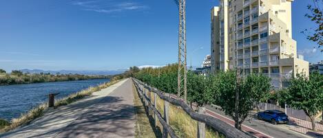 A riverside walkway with a wooden fence, a blue car, and a multi-story building with balconies.