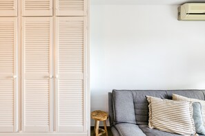 Een kamer met een grijze bank, een houten kruk en een witte kast met horizontale luiken.