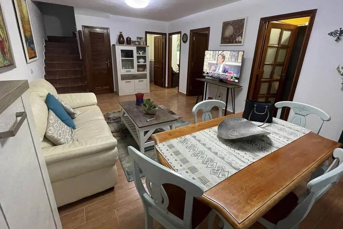 Una sala de estar con sofá, mesa de comedor, sillas, televisor y una escalera que conduce a otra habitación.