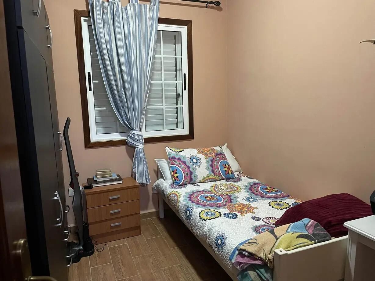 Un dormitorio pequeño con cama, mesita de noche, cómoda de madera, ventana con cortinas y puerta.