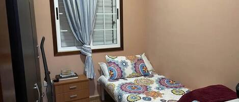 Un dormitorio pequeño con cama, mesita de noche, cómoda de madera, ventana con cortinas y puerta.