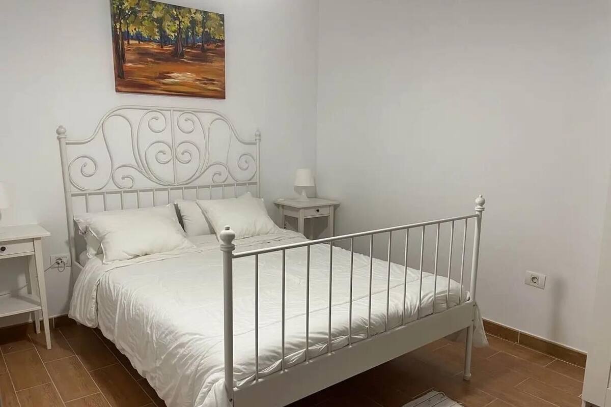 Una cama de metal blanca con cabecera, una mesita de noche, una lámpara y un cuadro en la pared.