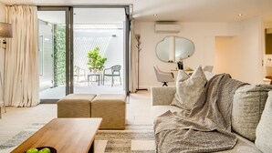 Ein modernes Wohnzimmer mit Sofa, Hocker und Couchtisch. Ein Schiebetürfenster führt auf einen Balkon mit Gartenmöbeln.