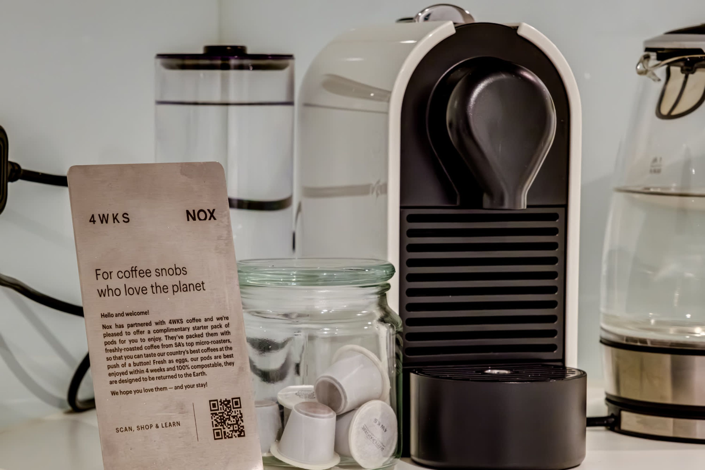 Eine Kaffeemaschine, ein Glasbehälter mit Kaffeekapseln und ein Schild mit Produktinformationen.