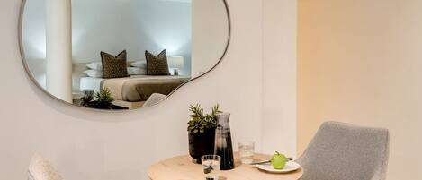 A modern room with a round table, two grey armchairs, a mirror, a bed with patterned pillows, a potted plant, and a green apple on a plate.