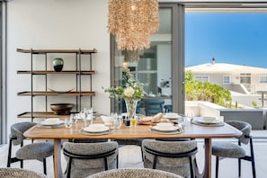 A dining area with a wooden table set for six, surrounded by upholstered chairs. A woven light fixture hangs above, and a vase with flowers is on the table. A metal shelf with a bowl and a vase is to the left. - Enjoy a Tranquil Holiday at Blue Ocean Residence (Cape Town)