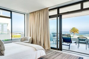 A modern hotel room with a large bed, a seating area with a view of the ocean, and a balcony with a palm tree. - Enjoy a Tranquil Holiday at Blue Ocean Residence (Cape Town)