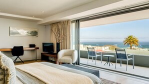 Una habitación de hotel moderna con una cama grande, un escritorio con computadora, televisor y balcón con mobiliario exterior.