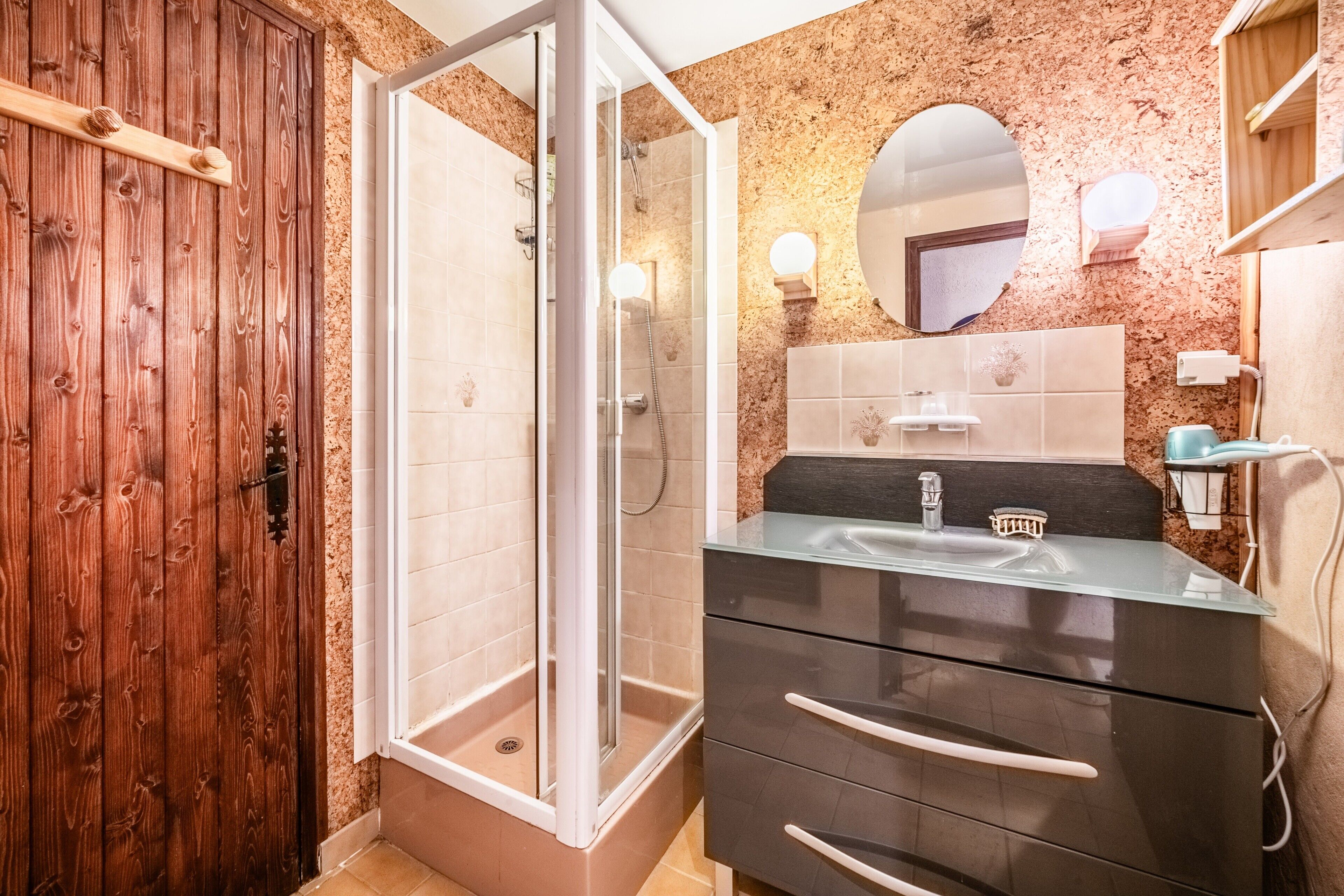 A bathroom with a wooden door, a glass shower enclosure, a black vanity with a glass top, and a wall-mounted mirror.